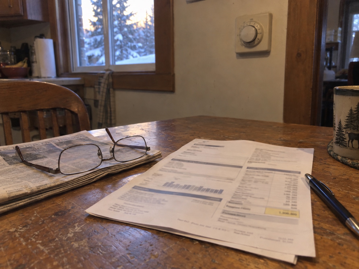 A heating bill, reading glasses, and a pen on a kitchen table, an old round thermostat on the wall, snow outside the window.